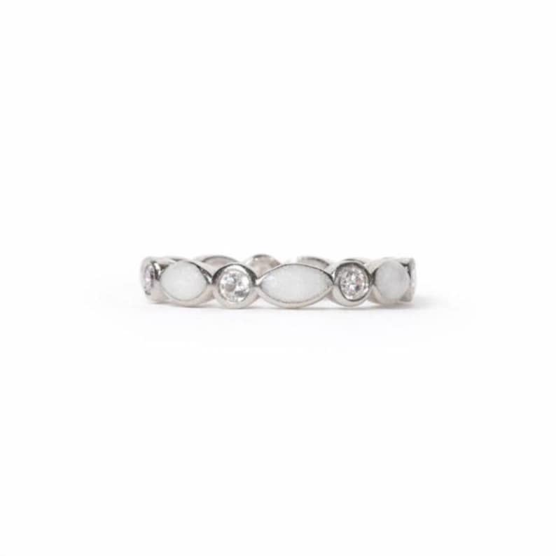 May include: A silver ring with a repeating pattern of white oval and round stones, alternating with clear, round gemstones. The ring is set against a white background, highlighting its delicate design and sparkle.