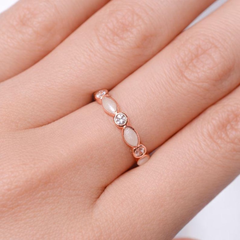 May include: Rose gold ring featuring alternating oval and round white stones, with a central clear, round stone. The ring is set on a finger, showcasing its delicate design and elegant style. A piece of jewelry.