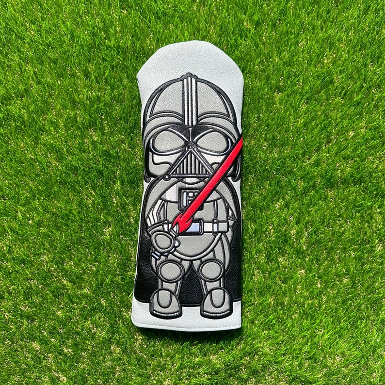May include: A golf club head cover featuring a cartoon depiction of Darth Vader. The cover is primarily black, gray, and white, with a red lightsaber. The design is detailed with a stitched appearance.