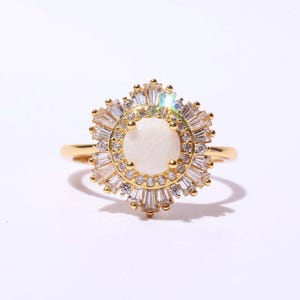 May include: A gold-toned ring featuring a central white stone surrounded by a halo of baguette-cut and round clear crystals. The ring's design resembles a flower or sunburst, with a delicate band.