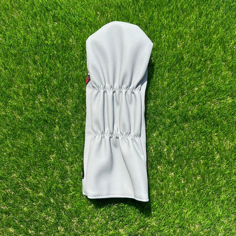 May include: A light gray golf club head cover made of textured material. The cover has a rounded top and a gathered design along the body. The cover is on a bed of green grass.