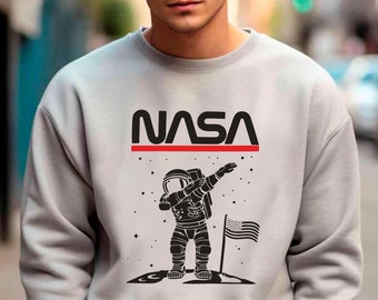 Peut inclure: Un sweat-shirt gris avec une illustration en noir et blanc d'un astronaute faisant le dab. L'astronaute se tient sur la lune avec un petit drapeau américain planté dans le sol. Le texte "NASA" est imprimé en noir au-dessus de l'astronaute avec une ligne rouge en dessous.