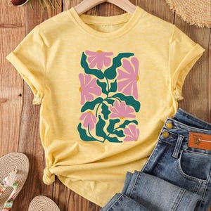May include: A yellow short-sleeved t-shirt with a floral design in pink and green. The shirt is on a wooden surface with a pair of sandals and a pair of jeans.