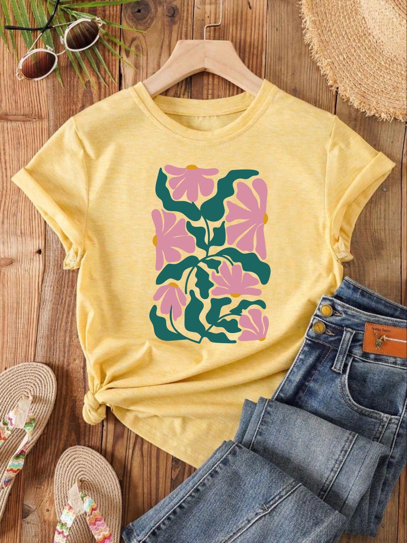 May include: A yellow short-sleeved t-shirt with a floral design in pink and green. The shirt is on a wooden surface with a pair of sandals and a pair of jeans.