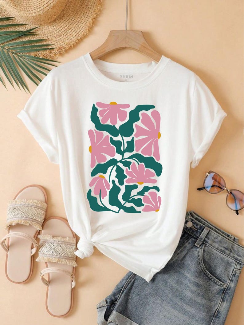 May include: White t-shirt with a pink and green floral print. The shirt has a crew neck and short sleeves. The print features stylized flowers and leaves.
