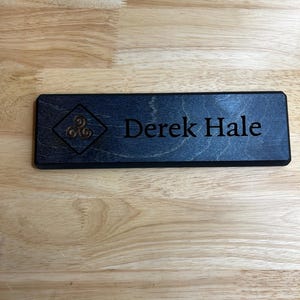 May include: A rectangular wooden sign with the name "Derek Hale" engraved in a dark font. The sign features a Celtic knot design in the upper left corner. The wood has a dark stain.