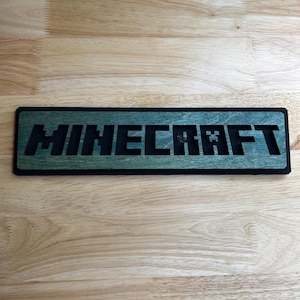 May include: A rectangular wooden sign with the word "MINECRAFT" cut out in black letters. The letters are set against a green background, with a black border. The sign is placed on a light brown wooden surface.