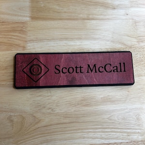 May include: A rectangular wooden name tag with the name "Scott McCall" engraved in a serif font. The tag has a dark red finish with a black border and a diamond-shaped graphic.