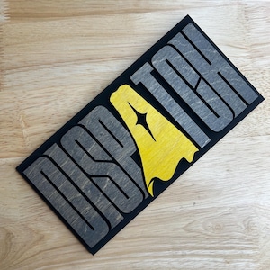 May include: A wooden sign with the word "DISPATCH" in gray, stylized letters. The letter "D" is replaced with a yellow shape with a star. The sign is on a black background.
