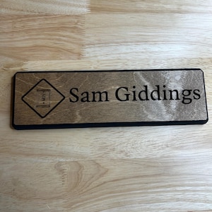 May include: A rectangular wooden nameplate with the name "Sam Giddings" in a black font. The nameplate has a black border and a diamond-shaped graphic with a door icon. The wood has a light brown color.