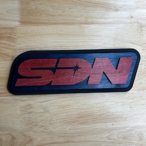 May include: Wooden sign with the letters "SDN" in red, set against a blue background with a black border. A starburst design is incorporated into the letter "D". The sign is placed on a wooden surface.