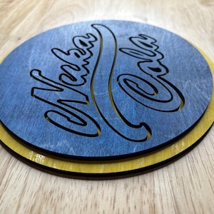 May include: A round wooden coaster with the words "Verba Cola" cut out in a script font. The top layer is blue, and the bottom layer is yellow. The coaster is sitting on a wooden surface.