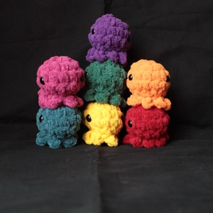 May include: A stack of seven crocheted octopus plushies in a rainbow of colors, including purple, pink, green, orange, yellow, red, and teal.