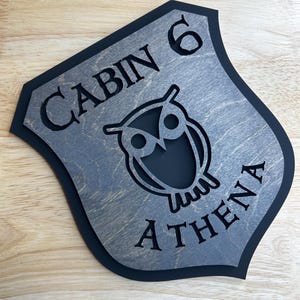 May include: Wooden sign with a shield shape, featuring the text "CABIN 6 ATHENA" and an owl silhouette. The sign has a gray wood grain finish with a black border, suitable for cabin decor.
