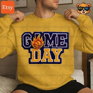 Puede incluir: Una sudadera amarilla con un diseño azul y amarillo de "GAME DAY". El diseño presenta una pelota de baloncesto con llamas. La sudadera es de estilo cuello redondo. El logotipo de Etsy está en la esquina superior izquierda.