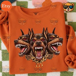 Puede incluir: Sudadera naranja con un gráfico de un perro de tres cabezas con caras amenazantes, ojos rojos y joyas doradas. El diseño está centrado en la parte delantera de la prenda de manga larga.