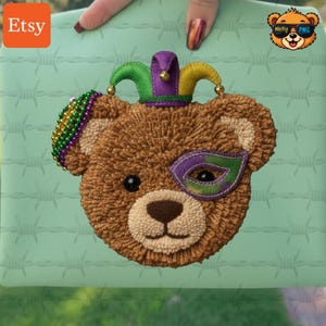 May include: A brown teddy bear face with a Mardi Gras theme, featuring a green, purple, and yellow jester hat and a matching eye mask. The Etsy logo is in the upper left corner, and a cartoon bear wearing sunglasses is in the upper right corner.