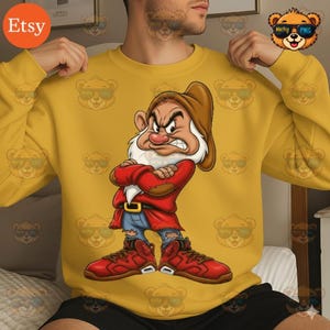 以下が含まれることがあります： 白雪姫のグランピーの漫画のイメージが描かれたマスタードイエローのスウェットシャツ。グランピーは厳しい表情で、赤いコート、青いジーンズ、赤いスニーカーを着用しています。 Etsyのロゴが見えます。