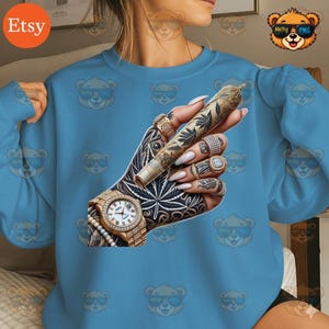 May include: Light blue sweatshirt featuring a detailed graphic of a hand with tattoos, rings, and a gold watch, holding a rolled cannabis cigarette. The hand is intricately tattooed with cannabis leaves and other designs.