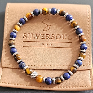 May include: A bracelet with blue lapis lazuli and tiger's eye beads, interspersed with silver-coloured metal spacers. The bracelet is displayed on a tan leather pouch, with the text "SILVERSOUL MEN".