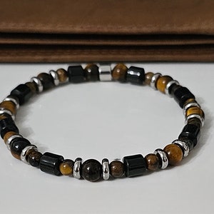 May include: A beaded bracelet featuring a mix of black, brown, and gold-colored beads. The bracelet includes cylindrical and round beads, with silver-colored spacers. The bracelet is displayed on a white surface with a brown leather wallet in the background.