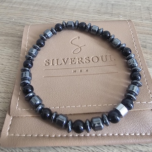 May include: A bracelet featuring a mix of black and silver beads. The bracelet sits on a tan leather pouch with the embossed text "SILVERSOUL MEN". The beads are a variety of shapes and sizes, creating a textured look.