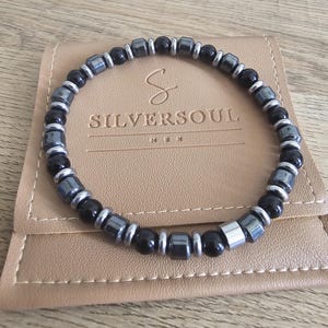 May include: A bracelet with black and silver beads is displayed on a tan leather pouch. The bracelet combines round black beads with rectangular silver-coloured beads. The pouch has the text "SILVERSOUL" printed on it.