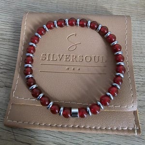Peut inclure: Un bracelet en perles rouges et argent avec un fermoir magnétique. Le bracelet est dans une pochette en cuir marron avec le texte "SILVERSOUL" imprimé dessus.