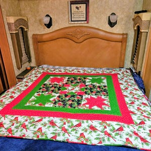 May include: A festive quilt adorns a bed, showcasing a Christmas theme. The quilt's design incorporates red, green, and white hues, with cardinal birds and star motifs. The bed is situated in a room with a wooden headboard and wall decorations.