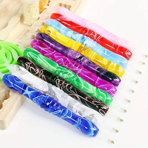 May include: A collection of colorful diamond painting pens with marbled designs. The pens are in various colors, including blue, green, purple, and pink. Each pen has a clear tip and is arranged on a wooden surface.