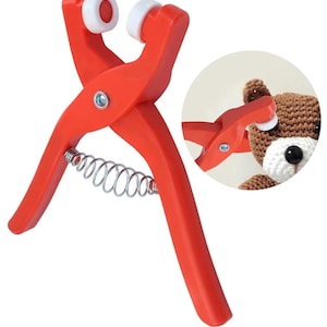 Op de afbeelding: Een rood knutselgereedschap met witte rollen en een zilveren veer. De tool is ontworpen om kleine objecten te bevestigen, zoals getoond in de inzetafbeelding met een gehaakte teddybeer.