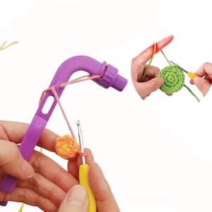 May include: A purple crochet tool with a curved handle and a pink yarn attached. The image also shows hands using a crochet hook to create a small orange and green crocheted circles. The background is white.