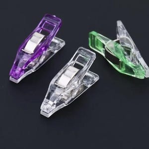 May include: Three clear plastic sewing clips in purple, green, and clear. Each clip has a silver metal hinge and a wide opening for holding fabric layers together. The clips are arranged on a dark background.