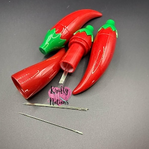 以下が含まれることがあります： 緑色のキャップが付いた赤い唐辛子型の針ホルダーと、いくつかの針。 ホルダーは開いており、内部の針が見えます。 画像には、ハート型のロゴで「Krafty Nations」というテキストも含まれています。