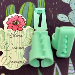 May include: Two light green plastic ring sizers with a textured surface. The sizers are open on one side and have a small hole on the other side. The sizers are designed to measure finger size.