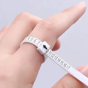 May include: A white ring sizer tool with black markings and numbers, used to measure finger size. The tool is being used on a finger, showing the measurement scale. The numbers range from 0 to 17.