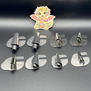 Könnte beinhalten: Ein Set aus acht silberfarbenen Metallwerkzeugen in verschiedenen Größen, von 3 mm bis 10 mm. Jedes Werkzeug hat eine einzigartige Form und ist für das Basteln konzipiert. Im Hintergrund befindet sich ein farbenfroher Drachenaufkleber.