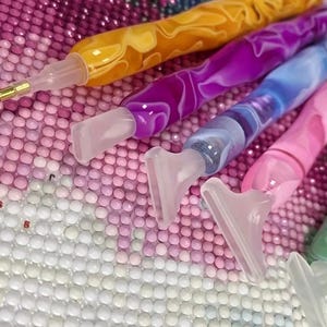 Puede incluir: Primer plano de varios bolígrafos de pintura de diamantes con mangos coloridos y veteados en tonos amarillos, morados, azules, rosas y verdes. Los bolígrafos tienen puntas claras y anguladas para recoger y colocar pedrería pequeña.