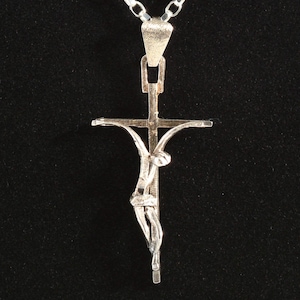May include: A silver crucifix pendant necklace. The pendant shows a detailed figure of Jesus Christ on the cross. The necklace has a simple chain and is set against a black background.