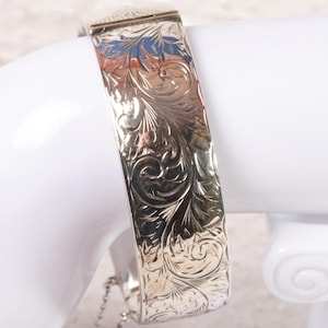 Peut inclure: Bracelet-jonc argenté avec un motif floral gravé. Le bracelet est présenté sur une surface blanche. La fermeture est sécurisée par une petite chaîne. Le design du bracelet présente des feuilles et des vignes détaillées et tourbillonnantes.
