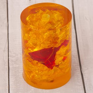 May include: A cylindrical, amber-coloured resin candle holder with a red accent. The candle holder is filled with gold-coloured, shredded material. The base of the candle holder is a darker amber colour.