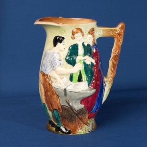 Op de afbeelding: Een vintage keramische kan met een gedetailleerde reliëfscène. De kan heeft een handvat dat is ontworpen om op een tak te lijken. De scène toont drie figuren, waarvan één aan een aambeeld werkt. De kan heeft een crèmekleurige achtergrond met bruine, groene, blauwe en rode accenten.