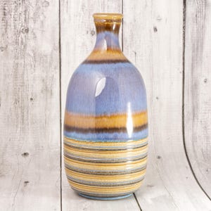 May include: A ceramic vase with a narrow neck and a rounded body. The upper portion features a smooth, glossy glaze in shades of blue and brown, while the lower section is decorated with numerous horizontal stripes in yellow, brown, and blue tones.