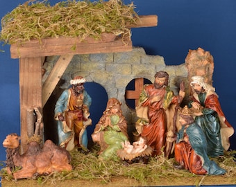 May include: A detailed nativity scene with figures of Mary, Joseph, baby Jesus, the three wise men, and a camel. The figures are set in a rustic stable with a stone wall and wooden beams, all on a bed of green moss.