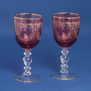 May include: Two matching wine glasses featuring deep purple bowls with intricate gold filigree floral patterns and gold rims. Each glass has a clear, multi-tiered decorative stem and a circular base with a thin gold edge detail.