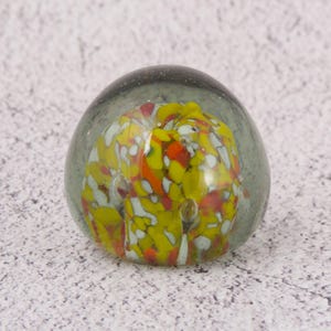 Könnte beinhalten: Ein klarer Glasbriefbeschwerer mit farbenfrohem Innenleben. Das Innere zeigt eine lebendige Mischung aus gelben, roten, orangen und weißen Glasfragmenten. Der Briefbeschwerer hat eine abgerundete, ovale Form und liegt auf einer hellgrauen Oberfläche.