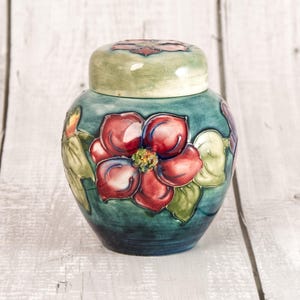 May include: A ceramic jar with a removable lid, featuring a deep teal blue base colour. The surface is decorated with large, raised relief red flowers and green leaves. The lid has a matching light green finish with a small red floral accent.