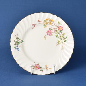 May include: A white porcelain plate with a fluted edge and a gold rim. The plate features a floral design with pink, yellow, and green flowers. The plate is displayed on a white stand against a blue backdrop.