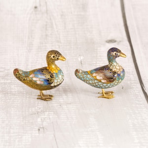 Puede incluir: Dos pequeñas figuras decorativas de patos elaboradas con un intrincado trabajo de esmalte cloisonné. Cada ave presenta un marco de metal dorado relleno con patrones vibrantes de escamas de esmalte azul, blanco, amarillo y marrón, sobre patas finas.