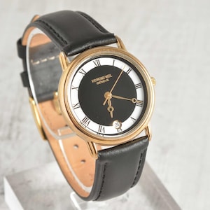 Può includere: Un orologio da polso color oro con quadrante nero e numeri romani. L'orologio ha un cinturino in pelle nera e il marchio "RAYMOND WEIL GENEVE" è stampato sul quadrante. L'orologio è esposto su un supporto bianco.